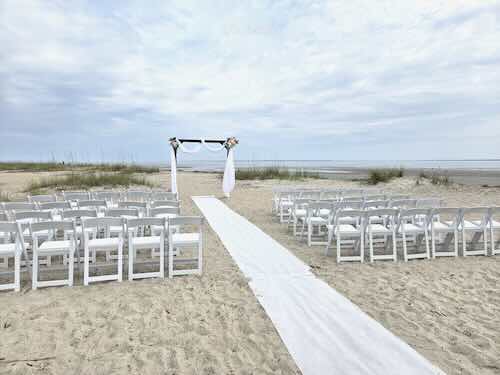 Beach wedding setup with white chairs and aisle.