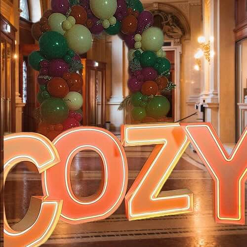 Balloon archway with glowing 'COZY' sign indoors.